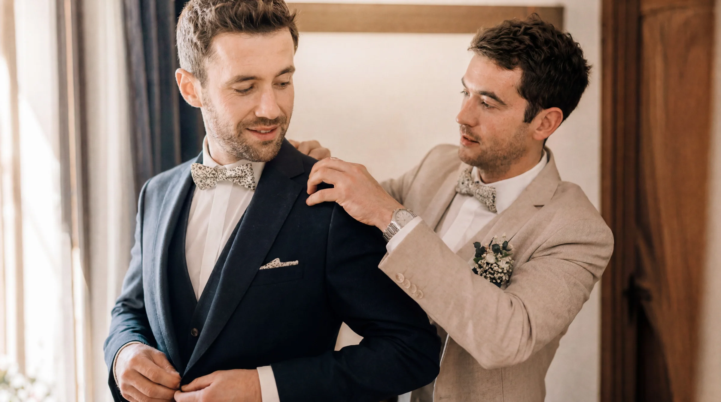 Un marié en costume bleu ajustant sa veste, aidé par un proche, lors d'un mariage à Valence, ambiance préparatifs