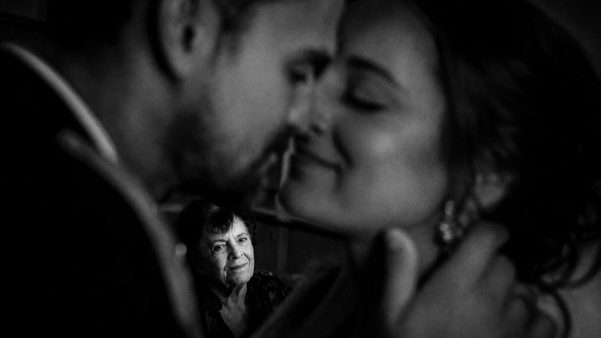 Reportage mariage à Romans, un couple s'embrasse tendrement sous le regard ému d'une invitée en noir et blanc